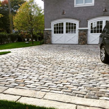 Cobble Paving Cobblestone driveway with a black car parked beside two garage doors.